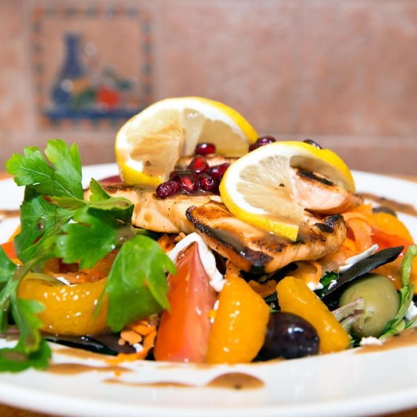 Grilled salmon salad with lemons, pomegranate, parsley, olives, and bell peppers on a white plate.