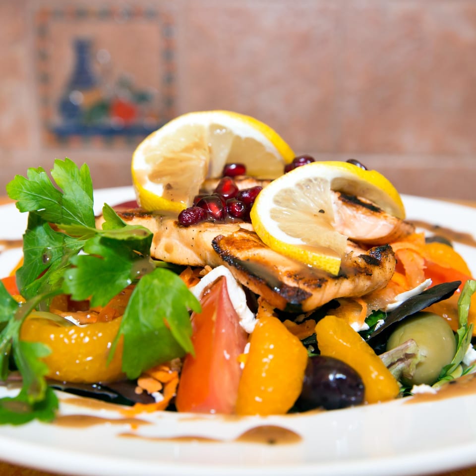 Grilled salmon salad with lemons, pomegranate, parsley, olives, and bell peppers on a white plate.