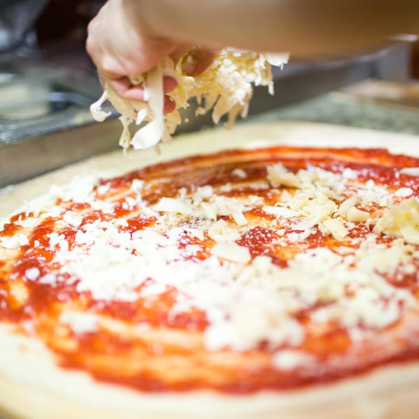 A hand sprinkles shredded cheese over a pizza crust with red sauce at Grandpa's Brick Oven Pizza.