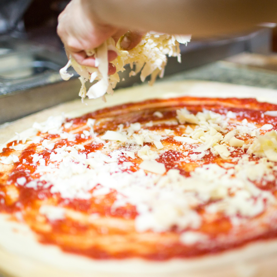 A hand sprinkles shredded cheese over a pizza crust with red sauce at Grandpa's Brick Oven Pizza.