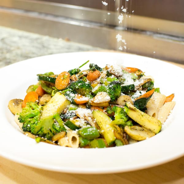 Pasta primavera with broccoli, carrots, zucchini, and mushrooms, topped with falling Parmesan cheese.
